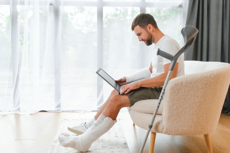 A man sitting on a couch with crutches and a laptop.