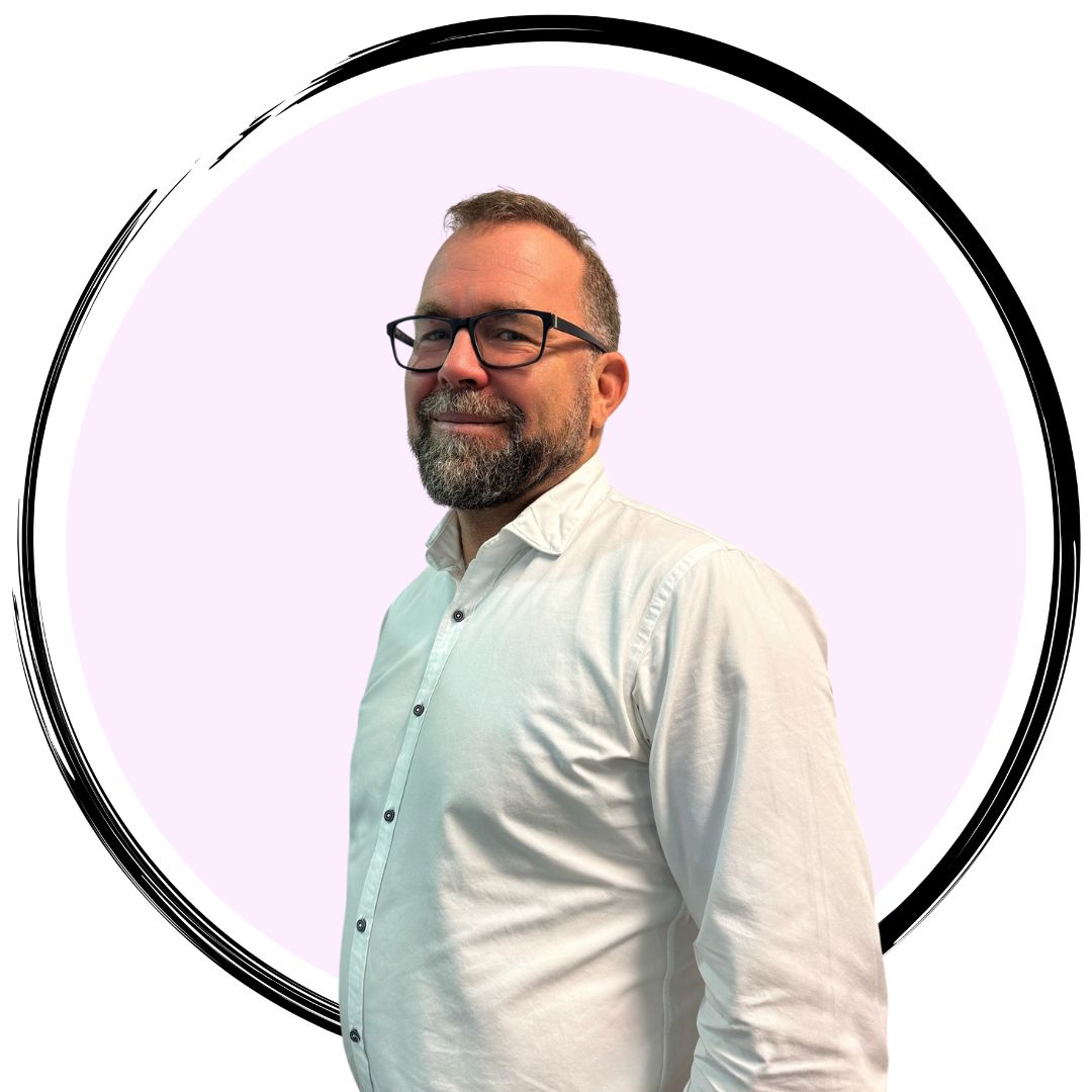 A man with a beard and glasses, wearing a white button-up shirt, is posing against a pink circular background with a black border. He stands slightly angled to the side, looking at the camera with a neutral expression.